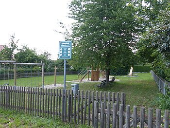 Spielplatz mit einem Holzzaun, Schaukeln, Klettergerüst, Rutsche, Bänken und Bäumen auf Grasfläche und teilweise mit Kieselsteinen bei Tageslicht. Außerdem ein am Zaun angebrachtes grünes Schild mit Regeln zur Spielplatzbenutzung und eine Mülltonne aus Metall zu sehen.