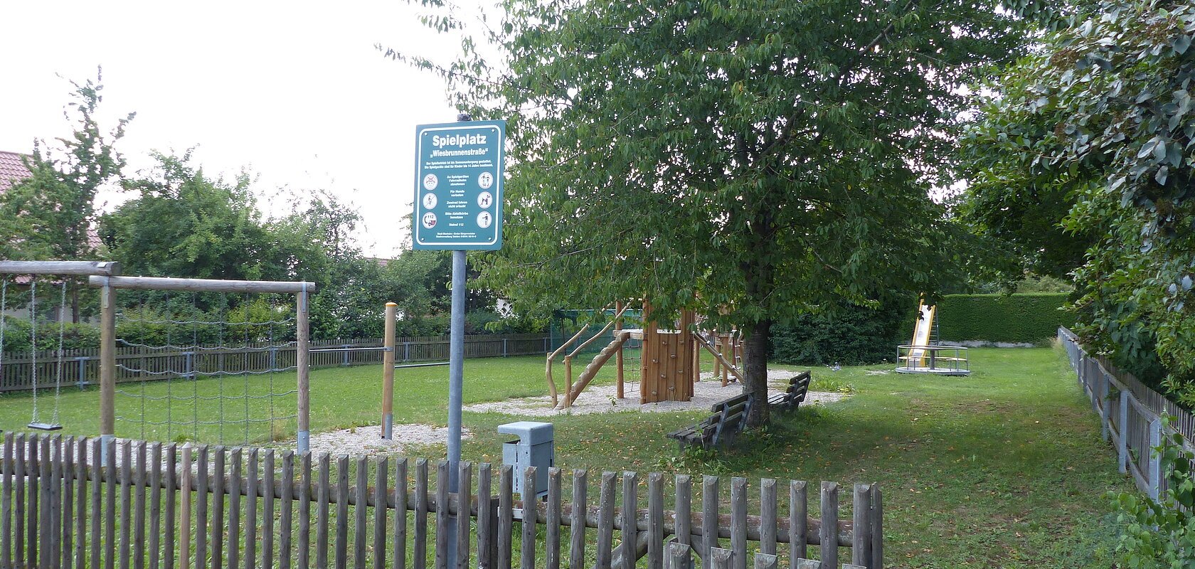 Spielplatz mit einem Holzzaun, Schaukeln, Klettergerüst, Rutsche, Bänken und Bäumen auf Grasfläche und teilweise mit Kieselsteinen bei Tageslicht. Außerdem ein am Zaun angebrachtes grünes Schild mit Regeln zur Spielplatzbenutzung und eine Mülltonne aus Metall zu sehen.