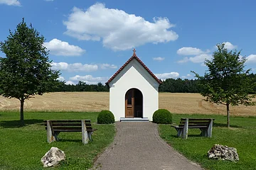 Kleine weiße Kapelle mit rotem Dach an einem gepflastertem Weg mit jeweils einer Bank, einem Baum und einem großen Stein links und rechts am Weg entlang, auf einer Wiese vor Feld und blauem Himmel.