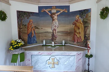Altar in einer Kapelle mit Kreuzigungsszene vom Jesus am Kreuz, Maria links, Johannes rechts an einer weißen Wand. Drei Kerzen stehen auf dem Altar und eine große Kerze rechts davon auf einem Kerzenständer. Blumenarrangement links am Altar und zwei grüne Kränze an der Wand angebracht.