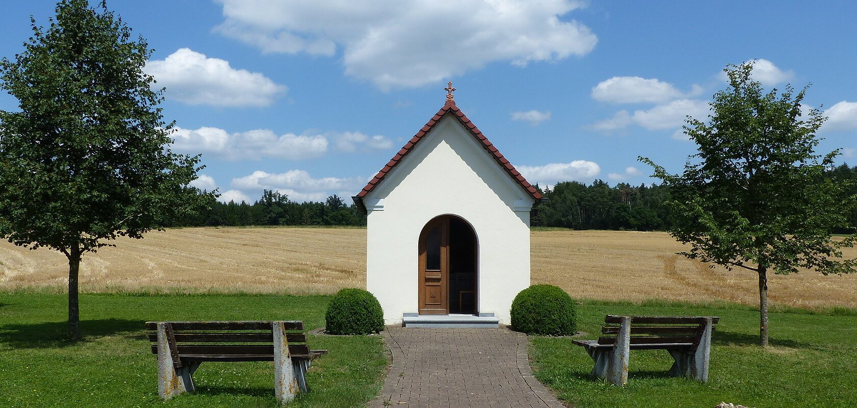 Kleine weiße Kapelle mit rotem Dach an einem gepflastertem Weg mit jeweils einer Bank, einem Baum und einem großen Stein links und rechts am Weg entlang, auf einer Wiese vor Feld und blauem Himmel.