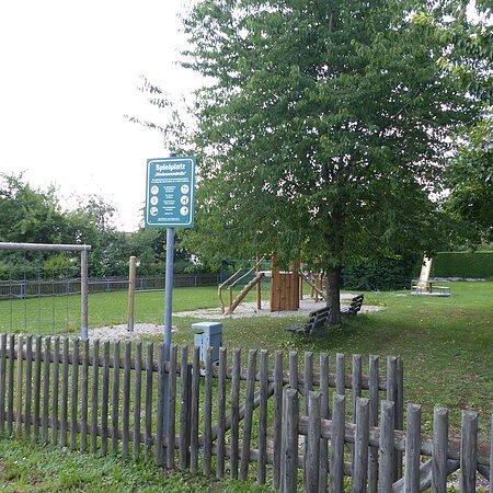 Spielplatz mit einem Holzzaun, Schaukeln, Klettergerüst, Rutsche, Bänken und Bäumen auf Grasfläche und teilweise mit Kieselsteinen bei Tageslicht. Außerdem ein am Zaun angebrachtes grünes Schild mit Regeln zur Spielplatzbenutzung und eine Mülltonne aus Metall zu sehen.