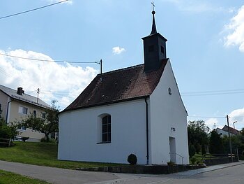 Kleine weiße Filialkirche mit rotem Ziegeldach und kleinem Turm oben drauf mit Kreuz an einer Straße bei klarem Himmel. Dahinter ein weiteres Wohnhaus mit grüner Wiese und Bäumen.