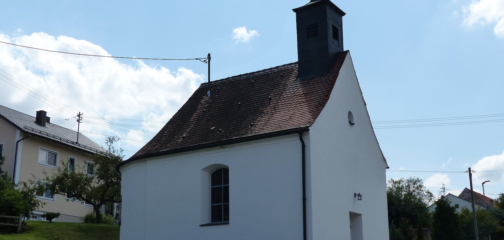 Kleine weiße Filialkirche mit rotem Ziegeldach und kleinem Turm oben drauf mit Kreuz an einer Straße bei klarem Himmel. Dahinter ein weiteres Wohnhaus mit grüner Wiese und Bäumen.
