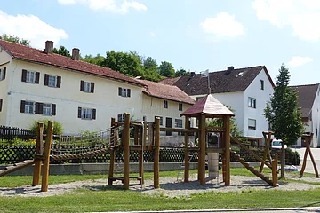 Spielplatz mit einem großen Klettergerüst aus Holz und einer Nestschaukel aus Holz auf grüner Wiese mit Kieselsteinen und einem Baum, vor Wohnhäusern und blauem Himmel am Tag.