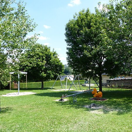 Spielplatz mit Wippen, Schaukeln und einem Sandkasten umgeben von grünen Bäumen auf einer Wiese bei Sonnenschein.