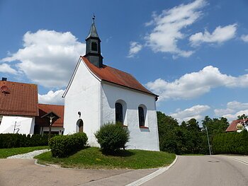 Kleine weiße Filialkirche mit rotem Dach und kleinem Turm oben drauf an einer Straße bei blauem Himmel mit Wolken. Umgeben von grüner Wiese und Hecken, daneben ein Jesuskreuz aus Holz. Im Hintergrund weiteres weißes Haus zu sehen.