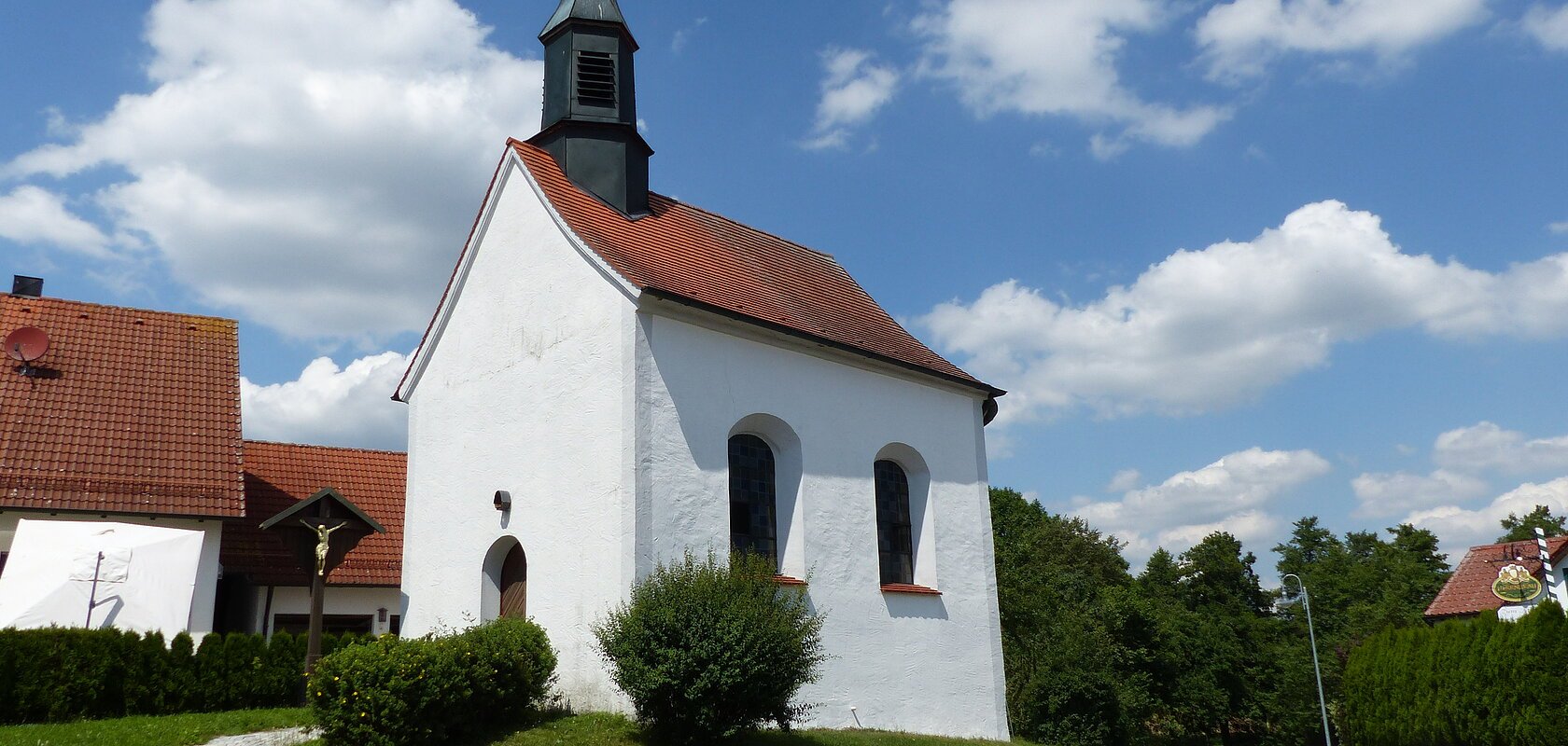 Kleine weiße Filialkirche mit rotem Dach und kleinem Turm oben drauf an einer Straße bei blauem Himmel mit Wolken. Umgeben von grüner Wiese und Hecken, daneben ein Jesuskreuz aus Holz. Im Hintergrund weiteres weißes Haus zu sehen.