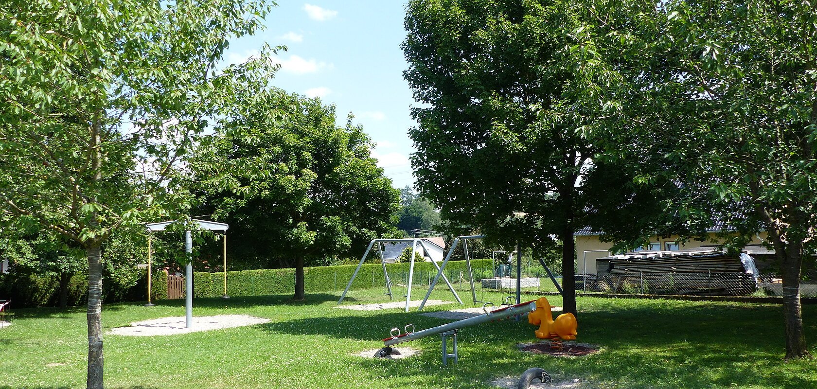 Spielplatz mit Wippen, Schaukeln und einem Sandkasten umgeben von grünen Bäumen auf einer Wiese bei Sonnenschein.
