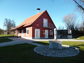 Rotes Gebäude mit Giebeldach, Fenstern, einem Balkon und einem Vordach. Im Vordergrund ein Brunnen auf gepflastertem Platz bei klarem Himmel. Um das Haus grüne Wiese und Bäume.
