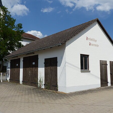 Weißes Gebäude mit braunem Dach und braunen Holztüren, darauf ein Schriftzug „Freiwillige Feuerwehr“ in braun/rot auf einem gepflastertem Platz. Im Hintergrund ein hoher Baum und eine Kirche mit Turm unter blauem Himmel zu sehen.
