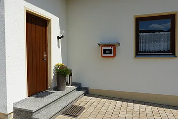 Eingangsbereich eines Hauses mit weißer Fassade, einer brauner Holztür, zwei Stufen davor, einem bepflanzten Blumentopf, einem Fenster mit Gardinen rechts im Bild und einem Defibrillator in einem orangen Kasten, der an der weißer Wand angebracht ist.