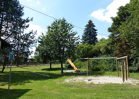 Großer Spielplatz mit einer Seilbahn, Schaukel, Rutsche, Wippe und Sandfläche, umgeben von Bäumen und Wiese bei Sonnenschein. Links im Bild steht ein Pfosten mit einem grünen Schild, auf dem die Spielregeln draufstehen.