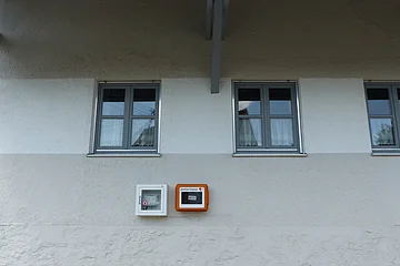 Fassade eines weißen Hauses mit drei Fenstern und zwei Kästen für Defibrillatoren, die an der Wand befestigt sind.