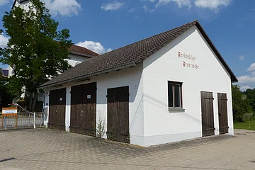 Weißes Gebäude mit braunem Dach und braunen Holztüren, darauf ein Schriftzug „Freiwillige Feuerwehr“ in braun/rot auf einem gepflastertem Platz. Im Hintergrund ein hoher Baum und eine Kirche mit Turm unter blauem Himmel zu sehen.