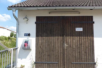 Braunes Holztor eines weißen Gebäudes mit weißem Schild drauf „Feuerwehr - Ausfahrt freihalten“ und automatischem externen Defibrillator an der Wand. Links im Bild im Hintergrund ein weißes Haus mit grüner Wiese bei blauem Himmel.