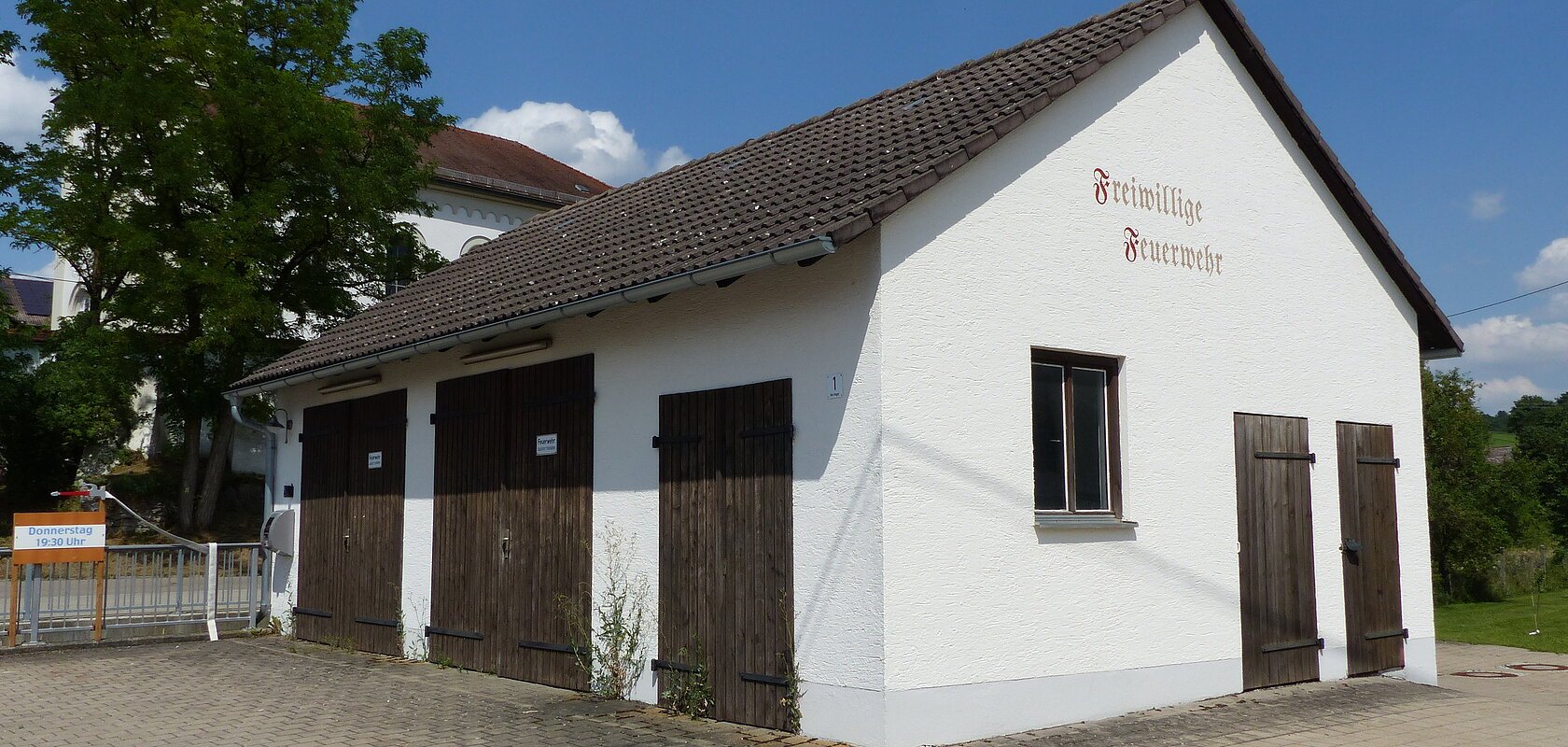 Weißes Gebäude mit braunem Dach und braunen Holztüren, darauf ein Schriftzug „Freiwillige Feuerwehr“ in braun/rot auf einem gepflastertem Platz. Im Hintergrund ein hoher Baum und eine Kirche mit Turm unter blauem Himmel zu sehen.