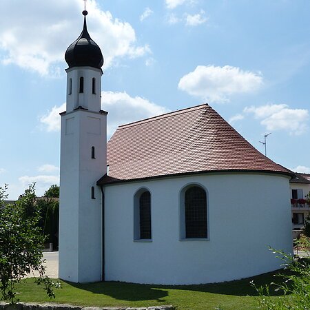 Weißes Kirchgebäude mit abgerundetem Grundriss und kleinem Turm mit schwarzer Haube bei blauem Himmel, umgeben von grünen Bäumen und wiese. Im Hintergrund weitere Häuser.