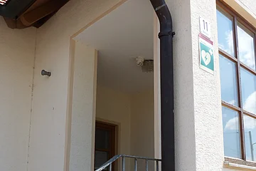 Eingang zu einem Feuerwehrhaus mit Treppe, Metallgeländer, Fenster und grünem Defibrillator-Schild an der Hauswand mit Hausnummer 11.
