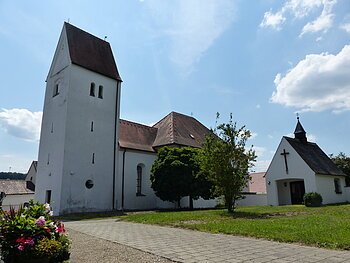 Filialkirche mit rotem Dach und einer kleiner Kapelle daneben mit einem Turm bei blauem Himmel und grünem Rasen im Vordergrund. Neben dem Rasen verläuft ein gepflasterter Gehweg mit Schottersteinen daneben.