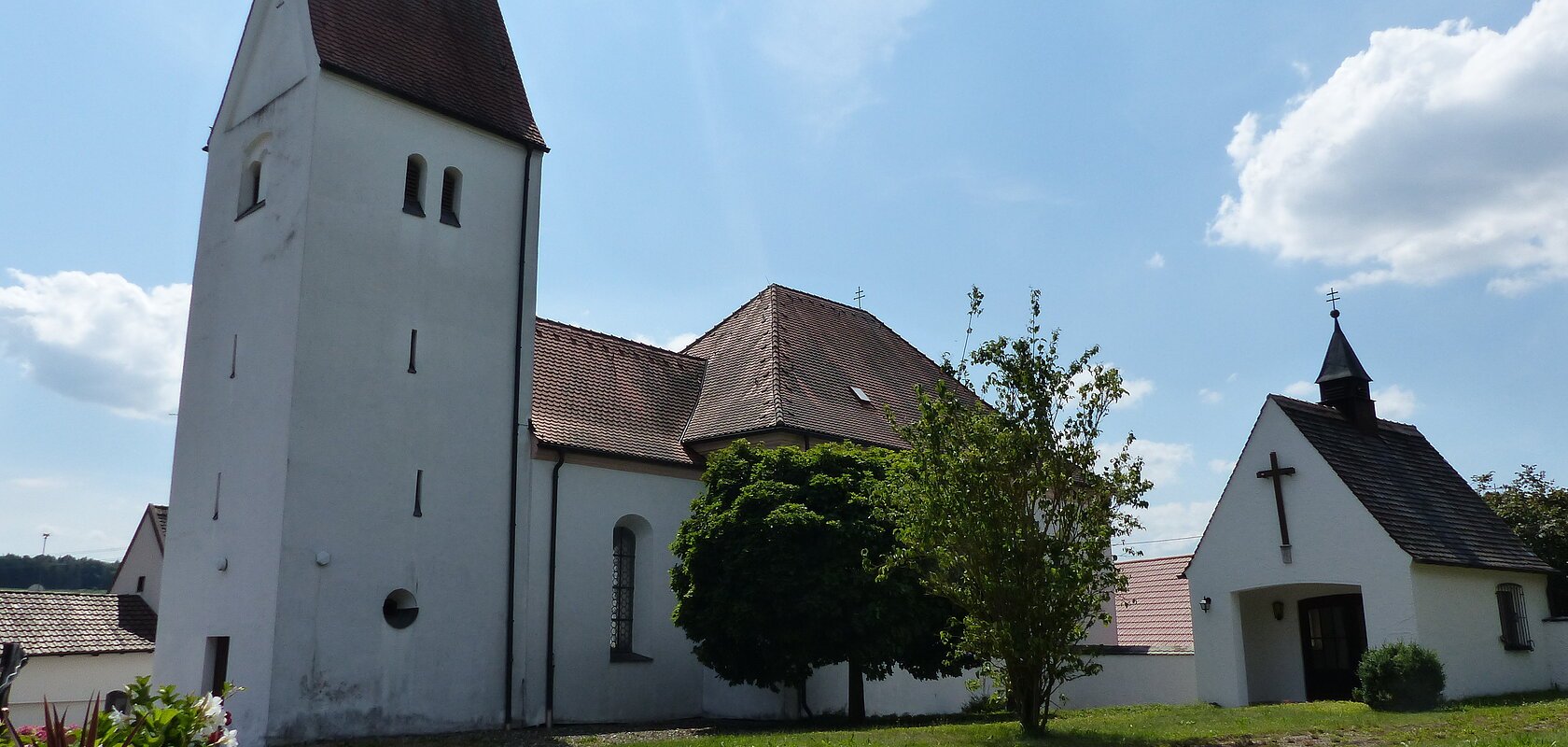Filialkirche mit rotem Dach und einer kleiner Kapelle daneben mit einem Turm bei blauem Himmel und grünem Rasen im Vordergrund. Neben dem Rasen verläuft ein gepflasterter Gehweg mit Schottersteinen daneben.