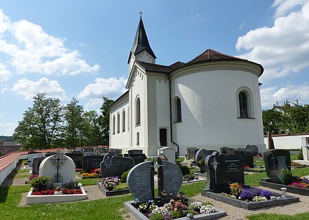 Weiße Pfarrkirche mit abgerundetem Gebäude, dunklem Dach und einem kleinen Turm mit schwarzem Dach. Anliegend ein Friedhof  mit Grabbepflanzung und Grabsteinen bei sonnigem Himmel und wenigen Wolken.