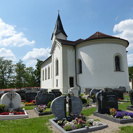 Weiße Pfarrkirche mit abgerundetem Gebäude, dunklem Dach und einem kleinen Turm mit schwarzem Dach. Anliegend ein Friedhof  mit Grabbepflanzung und Grabsteinen bei sonnigem Himmel und wenigen Wolken.