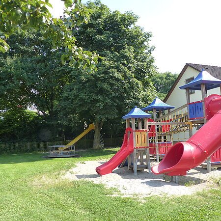 Spielplatz mit zusammenhängenden Spielgeräten wie zwei roten Rutschen, Klettergerüst und Sandfläche unten, neben einem weißen Wohnhaus. Im Hintergrund eine weitere gelbe Rutsche mit einem Karussell umgeben von Bäumen. Links im Bild teilweise ein grünes Fußballtor zu sehen.