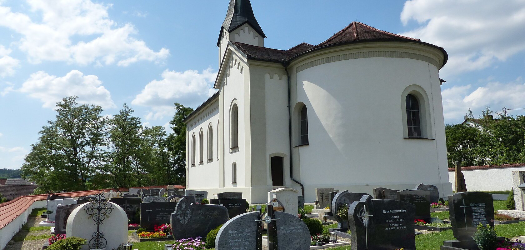 Weiße Pfarrkirche mit abgerundetem Gebäude, dunklem Dach und einem kleinen Turm mit schwarzem Dach. Anliegend ein Friedhof  mit Grabbepflanzung und Grabsteinen bei sonnigem Himmel und wenigen Wolken.