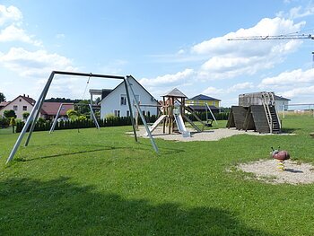 Spielplatz mit Nestschaukel, Rutsche aus Metall, Klettergerüst, Seilbahn und Wipp-Tier in Form eines Marienkäfers, auf einer Grasfläche. Im Hintergrund sind Wohnhäuser bei sonnigem Himmel. Rechts im Bild ein Teil eines Krans zu sehen.