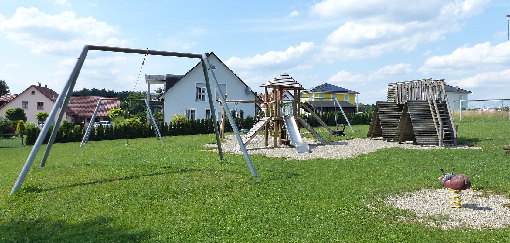 Spielplatz mit Nestschaukel, Rutsche aus Metall, Klettergerüst, Seilbahn und Wipp-Tier in Form eines Marienkäfers, auf einer Grasfläche. Im Hintergrund sind Wohnhäuser bei sonnigem Himmel. Rechts im Bild ein Teil eines Krans zu sehen.