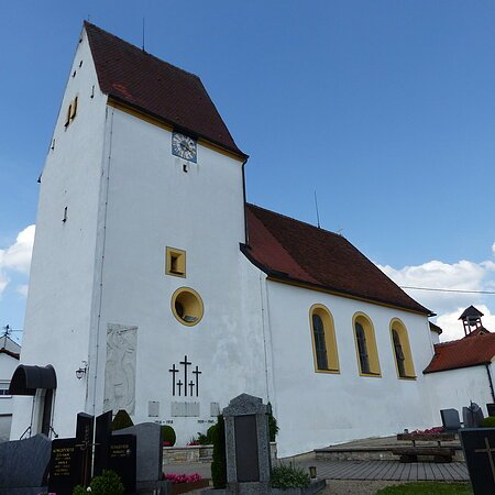 Weiße Pfarrkirche mit Turm, gelb eingerahmten Fenstern, einer Uhr und einem anliegenden Friedhof mit Grabsteinen, unter blauem Himmel.