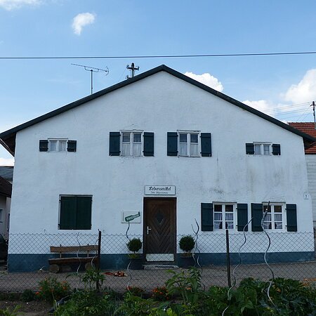 Weißes Haus mit grünen Fensterläden, einer braunen Holztür in der Mitte und einem weißen Schild darüber mit dem Schriftzug „Lebensmittel“. Im Vordergrund ist ein bepflanzter Garten mit einem Drahtzaun. Im Hintergrund weitere Häuser links und rechts und blauer Himmel.