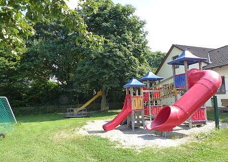 Spielplatz mit zusammenhängenden Spielgeräten wie zwei roten Rutschen, Klettergerüst und Sandfläche unten, neben einem weißen Wohnhaus. Im Hintergrund eine weitere gelbe Rutsche mit einem Karussell umgeben von Bäumen. Links im Bild teilweise ein grünes Fußballtor zu sehen.