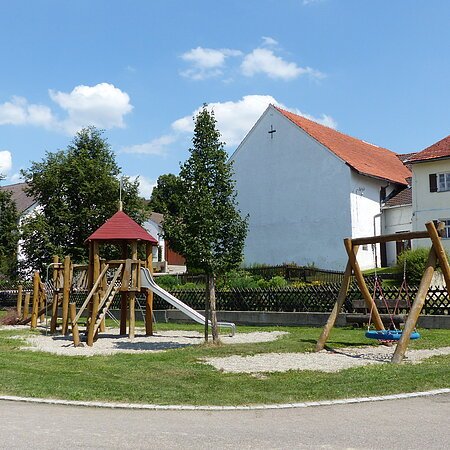 An einer Straße gelegener Spielplatz mit Klettergerüst, Rutsche und Nestschaukel. Umgeben von Bäumen, einer grünen Wiese, teilweise mit Kieselsteinen. Im Hintergrund weiße Häuser mit roten Dächern und blauem Himmel.
