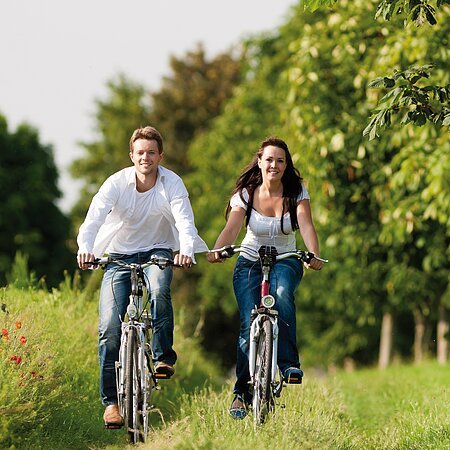 Zwei lächelnde Personen fahren nebeneinander auf Fahrrädern auf einem grasbewachsenen Weg mit Wiese links im Bild und Bäumen im Hintergrund.