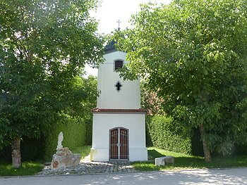 Kleine weiße Marienkapelle mit Fenster und einem Eingang, umgeben von Bäumen und Grünflächen an einer Straße. Vor der Kapelle links ein kleiner Brunnen aus Stein und ein großer Stein rechts als Sitzgelegenheit.