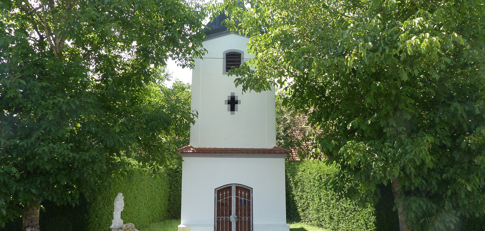 Kleine weiße Marienkapelle mit Fenster und einem Eingang, umgeben von Bäumen und Grünflächen an einer Straße. Vor der Kapelle links ein kleiner Brunnen aus Stein und ein großer Stein rechts als Sitzgelegenheit.