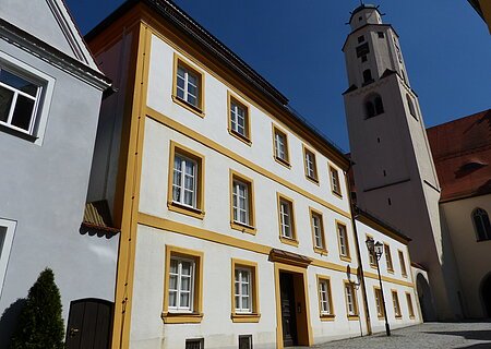 Dreistöckiges weißes Gebäude mit gelben Fensterrahmen und angrenzendem Kirchturm auf einem gepflastertem Platz, unter blauem Himmel.