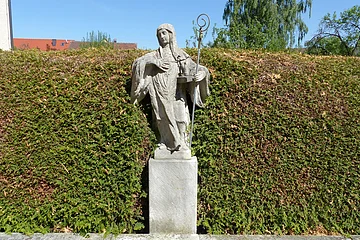 Steinerne Statue der Heiligen Walburga mit Stab vor einer hohen Hecke und blauem Himmel.