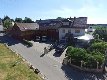 Haus mit großem Holz-Anbau und mit einem braunem Dach, geparkten Autos im Hof und einem grünen Garten mit Vordach, an einer Straße bei klarem Himmel.