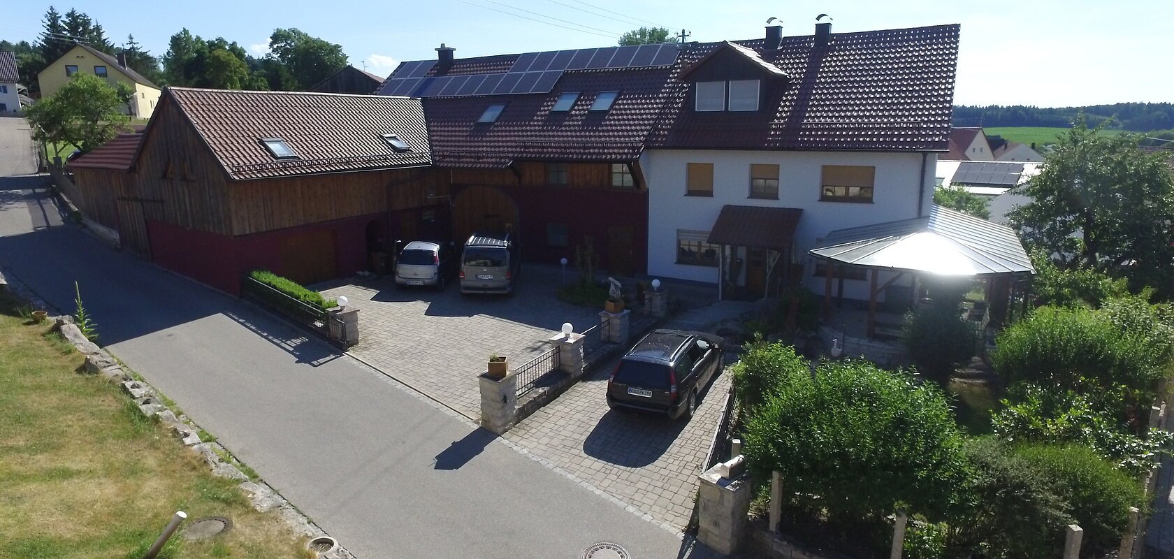 Haus mit großem Holz-Anbau und mit einem braunem Dach, geparkten Autos im Hof und einem grünen Garten mit Vordach, an einer Straße bei klarem Himmel.