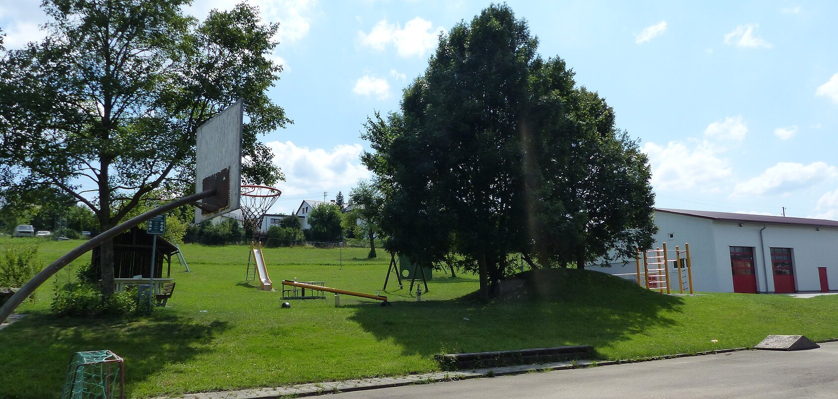 Spielplatz mit einem anliegenden Basketballkorb und einem kleiner Fußballtor darunter im Vordergrund. Im Hintergrund Spielplatz mit Rasenfläche mit Spielplatzgeräten wie Rutsche, Wippe, Klettergerüst, Schaukel und einem Trampolin umgeben von Bäumen bei sonnigem Wetter. Rechts daneben im Bild ein weißes Gebäude mit roten Toren.