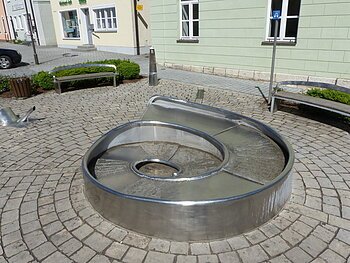 Juraschneck-Brunnen: ein Brunnen aus Metall in Form eines Fossils einer Schnecke, mit eingravierten Texten auf der Oberfläche. Außenrum ein gepflastertem Platz mit zwei Sitzbänken. Im Hintergrund farbige Häuser und eine kleine grüne Hecke an den Bänken entlang.