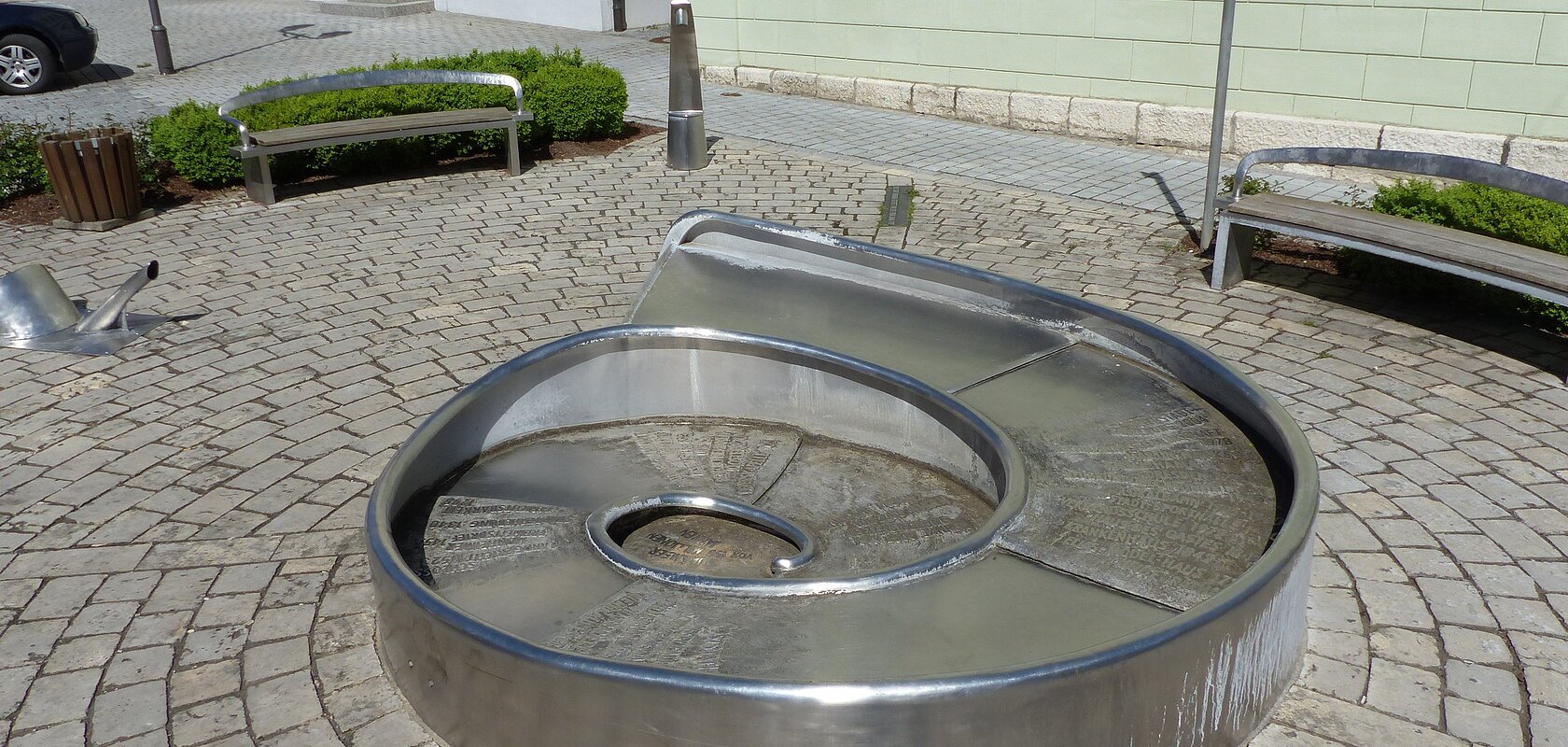Juraschneck-Brunnen: ein Brunnen aus Metall in Form eines Fossils einer Schnecke, mit eingravierten Texten auf der Oberfläche. Außenrum ein gepflastertem Platz mit zwei Sitzbänken. Im Hintergrund farbige Häuser und eine kleine grüne Hecke an den Bänken entlang.