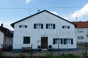 Weißes Haus mit grünen Fensterläden, einer braunen Holztür in der Mitte und einem weißen Schild darüber mit dem Schriftzug „Lebensmittel“. Im Vordergrund ist ein bepflanzter Garten mit einem Drahtzaun. Im Hintergrund weitere Häuser links und rechts und blauer Himmel.