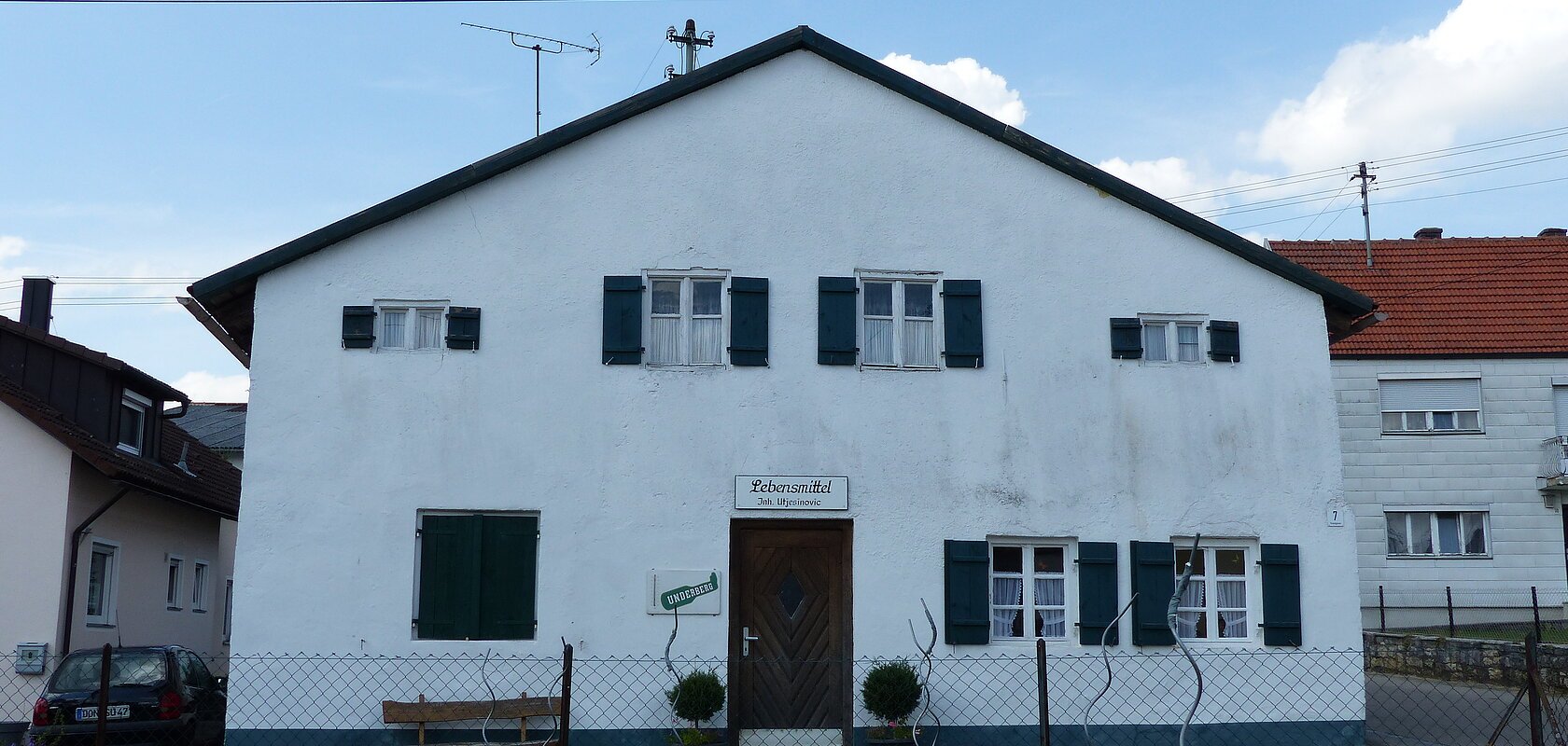 Weißes Haus mit grünen Fensterläden, einer braunen Holztür in der Mitte und einem weißen Schild darüber mit dem Schriftzug „Lebensmittel“. Im Vordergrund ist ein bepflanzter Garten mit einem Drahtzaun. Im Hintergrund weitere Häuser links und rechts und blauer Himmel.