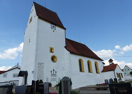 Weiße Pfarrkirche mit Turm, gelb eingerahmten Fenstern, einer Uhr und einem anliegenden Friedhof mit Grabsteinen, unter blauem Himmel.