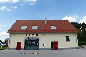 Gelbes Feuerwehrhaus mit rotem Satteldach, drei Dachfenstern und zwei Eingängen und einem Tor. Die Eingangstüren sind beide rot. Um das Gebäude große asphaltierte Fläche. Im Hintergrund blauer Himmel mit Bäumen und einem parkendem Auto.