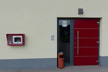 Gelbe Fassade eines Hauses mit roter Eingangstür mit Glasfenster. Links davon hängt ein roter Defibrillator-Kasten an der Wand und fein hoher roter Mülleimer steht vor der Haustür.